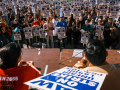 In the foreground we see the backs of a few strikers in red UAW 2865 T-shirts, one holding a strike sign, sitting at a table. Facing them is a huge crowd of strikers, standing, with picket signs, outdoors. The signs say "UAW on strike, unfair labor practice."