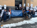 Postal workers rally outside a post office. There is some snow on the ground. A big blue banner says "Staffing, Safety, and Service: Letter Carriers Need a Raise! NALC Branch 9, Minneapolis." Printed picket signs say "End Mandatory Overtime," "First Class Service, First Class Pay," and "Fair Contract Now."