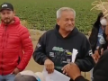 A masked worker in a straw hat speaks while an older unmasked man in a sweatshirt, the grower, listens, smiling or grimacing. Both are holding paper. A younger man in a red jacket, unmasked, stands impassive. Other masked people circle them, one with resolutely crossed arms.