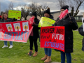 People stand on grass with signs. A red banner says "Seven months without a contract, OMG!" with stylized images of Edward Munch's painting "The Scream." Another sign says "More sick leave" in English, Spanish, and Russian. Another says, in Spanish,"Provide money for the future of students." Another says "Woodburn teachers don't want to strike, but we will." 