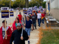 A dense line of picketers (at least 25 people are visible but it looks like there are more behind them) marches towards the camera on a leafy sidewalk alongside a factory. Many carry printed signs like "UAW on strike" and "Stand up for retirement security." Most visible faces are white but a few Black faces are also visible; there's a mix of women and men.