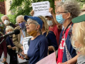 A group of older people in masks stands outside. The person in the middle is speaking into a mic. Someone behind her holds up a printed sign: "City Retirees Say NO, Don't Privatize Our Senior Care!"