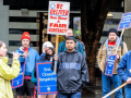 Letter carriers stand outdoors in the rain, rallying, listening to a speaker with a mic. Printed picket signs say "We deliver--how about a fair contract?" "End closed bargaining" "Harder work deserves a fair contract" and "Fair contract now."