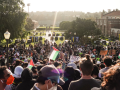 A large crowd, mostly viewed from behind, is arrayed on a hillside or steps of a university campus; banners identify it as UCLA. A few in the crowd hold Palestinian flags and one handmade sign reads "Stay strong." A row of parked vehicles is visible in the distance, at the foot of the hill, backs to the crowd. Dramatic sunlight silhouettes everyone.