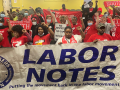 People stand indoors behind a big Labor Notes banner. Most are wearing red shirts and have their fists in the air. Several farther back are holding up smaller red "Organize the South" banners.