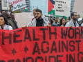 Workers stand outdoors, many wearing keffiyehs, holding a large red hand-painted banner that says "Health care workers against genocide in Gaza." Signs visible in crowd behind them say "Hospitals are not targets," "Free Palestine," and something ending in "slaughter." 