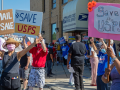 A group of people, masked, socially distanced, and diverse in race and gender, stand outside the Old Bedford Station post office on a sunny day. They hold up handmade signs: "US Mail Not 4 Sale" and "Save USPS." Someone in the back has a camera set up on a tripod. Similar printed signs from APWU are also visible.