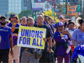 Workers march through a Detroit street, part of a Labor Day march. The people visible are mostly wearing purple SEIU T-shirts, many are Black and one holds a big printed sign readgin "Unions for all" on a yellow background. Another printed sign says "Families first, not billionaires. Not one more cut." One woman in a "Michigan home care workers" shirt pushes another in a wheelchair, also in purple gear.