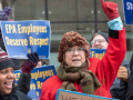 A crowd of federal workers stand outside, bundled against cold, carrying printed AFGE signs that say "EPA employees deserve respect" and "Stop the shutdown." In the foreground are a Black woman and a white woman, the latter raising a red-gloved fist in the air, both chanting or singing.