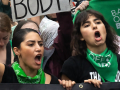 Two women in green bandanas that say “bans off our bodies” march with many others