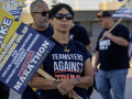 A woman with a strike picket sign and a ‘Teamsters Against Trump’ T-shirt looks past the camera