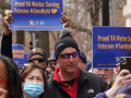 several people are jammed together as part of a march with blue signs with gold lettering, saying “Proud VA Worker Serving Veterans, #SaveMyVA”